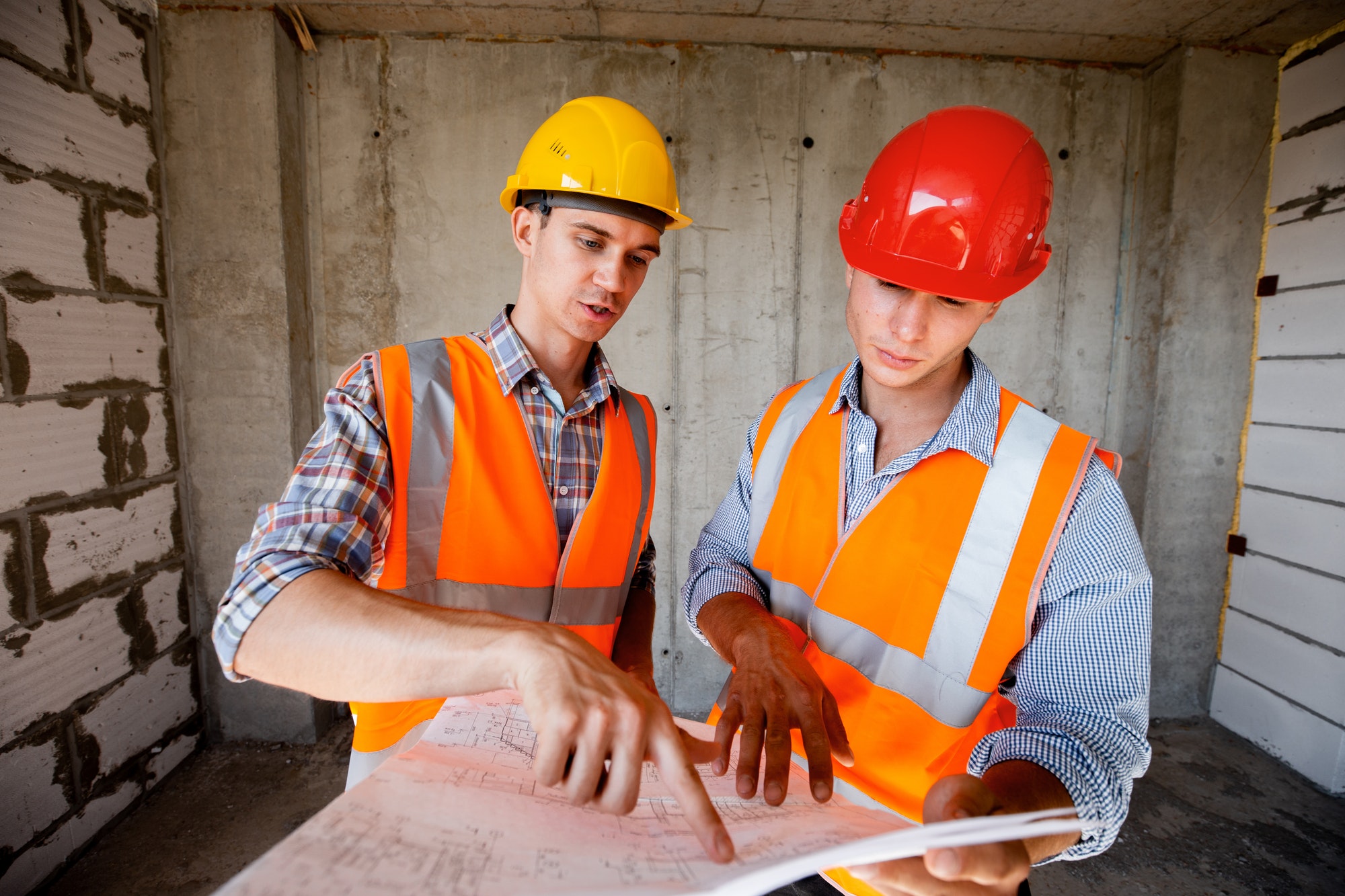 Two men dressed in orange work vests and helmets explores construction documentation on a concrete